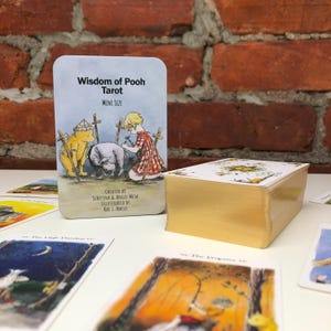 May include: A deck of "Wisdom of Pooh Tarot" cards, mini size, with gold gilded edges. The card art features illustrations of Winnie the Pooh, Piglet, and Christopher Robin. The deck is on a white surface with a brick background.