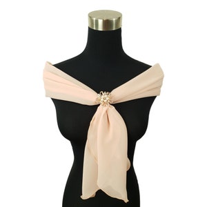 May include: A blush-colored chiffon shawl draped over a black mannequin. The shawl is secured with a gold-toned brooch featuring pearls and a floral design. The fabric is lightweight and flows gracefully, creating an elegant look.