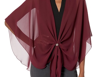 May include: A burgundy chiffon shawl, draped over a black top and fastened with a decorative clasp. The lightweight shawl is gathered and knotted at the front. The model is wearing grey trousers.