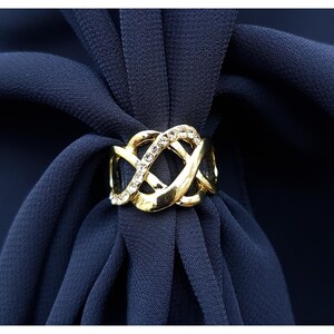 Puede incluir: Un anillo de bufanda dorado con un dise&ntilde;o entrelazado adornado con pedrer&iacute;a transparente. El anillo se coloca sobre un fondo de tela azul marino, creando un contraste llamativo. El anillo de bufanda a&ntilde;ade un toque de elegancia.