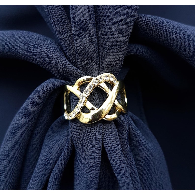 Puede incluir: Un anillo de bufanda dorado con un dise&ntilde;o entrelazado adornado con pedrer&iacute;a transparente. El anillo se coloca sobre un fondo de tela azul marino, creando un contraste llamativo. El anillo de bufanda a&ntilde;ade un toque de elegancia.