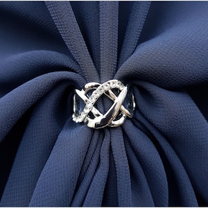 Puede incluir: Un anillo de bufanda plateado con un dise&ntilde;o intrincado e entrelazado, adornado con peque&ntilde;os cristales transparentes. El anillo se coloca sobre un fondo de tela azul oscuro, creando un contraste llamativo. Este accesorio a&ntilde;ade un toque de elegancia.