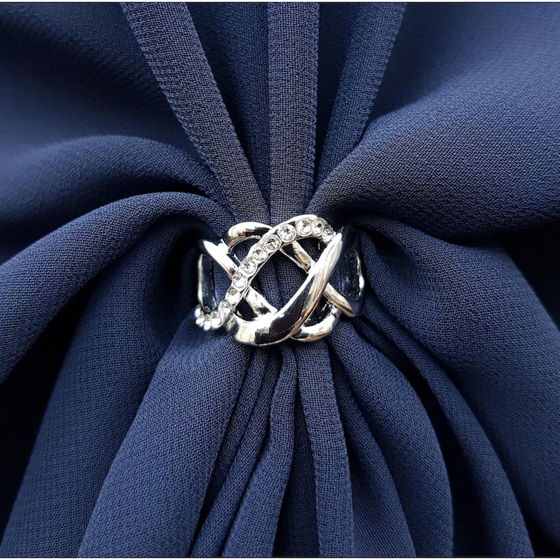 Puede incluir: Un anillo de bufanda plateado con un dise&ntilde;o intrincado e entrelazado, adornado con peque&ntilde;os cristales transparentes. El anillo se coloca sobre un fondo de tela azul oscuro, creando un contraste llamativo. Este accesorio a&ntilde;ade un toque de elegancia.