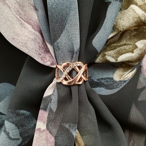 May include: A rose gold scarf ring with an intricate, intertwined design, set with clear crystals. The ring fastens a floral patterned scarf in grey, purple, and green. This accessory adds a touch of elegance to any outfit.