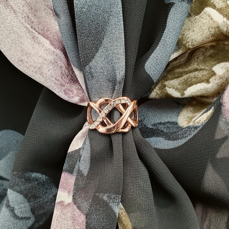 May include: A rose gold scarf ring with an intricate, intertwined design, set with clear crystals. The ring fastens a floral patterned scarf in grey, purple, and green. This accessory adds a touch of elegance to any outfit.
