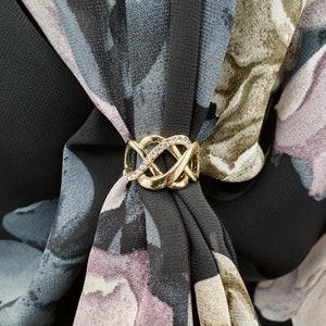 May include: A gold scarf ring with an intricate, interwoven design, set with clear rhinestones. The ring fastens a black scarf featuring a floral pattern in grey, purple, and olive green. The scarf is made of a lightweight material.