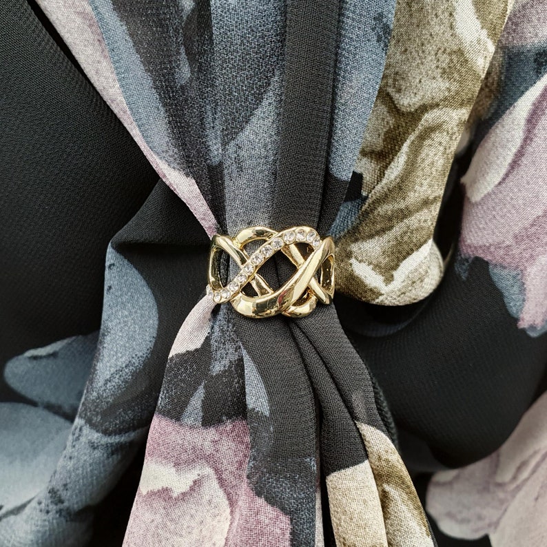 May include: A gold scarf ring with an intricate, interwoven design, set with clear rhinestones. The ring fastens a black scarf featuring a floral pattern in grey, purple, and olive green. The scarf is made of a lightweight material.