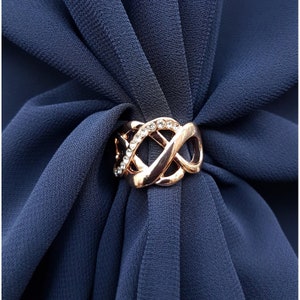 Puede incluir: Un anillo de bufanda de oro rosa con un dise&ntilde;o tejido y una fila de cristales transparentes. El anillo sujeta una bufanda azul marino. La bufanda tiene una textura suave y fluida.