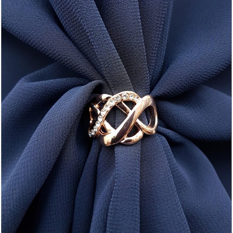 Puede incluir: Un anillo de bufanda de oro rosa con un dise&ntilde;o tejido y una fila de cristales transparentes. El anillo sujeta una bufanda azul marino. La bufanda tiene una textura suave y fluida.
