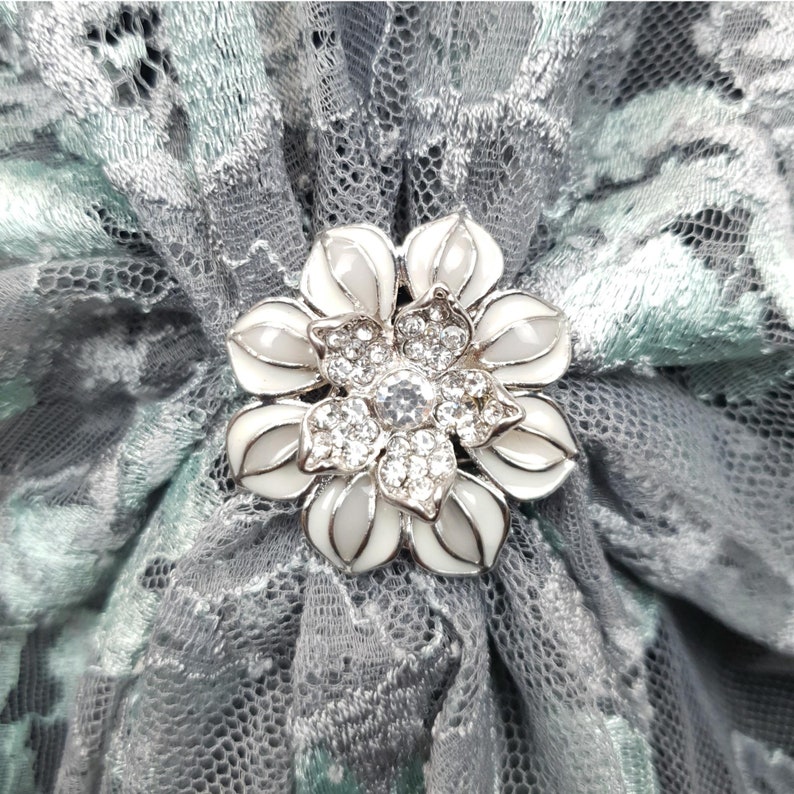 K&ouml;nnte beinhalten: Eine silberne Brosche in Blumenform mit wei&szlig;en Emaillebl&auml;ttern und einem Zentrum aus klaren Strasssteinen. Die Brosche befindet sich auf einem grauen und gr&uuml;nen Spitzenstoff.