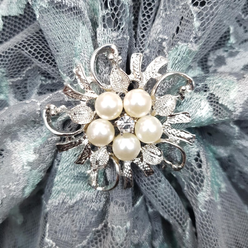 K&ouml;nnte beinhalten: Eine silberne Brosche mit floralem Design und f&uuml;nf Perlenimitationen. Die Brosche befindet sich in der Mitte eines hellblauen und grauen Spitzenstoffes.