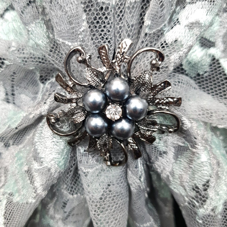 K&ouml;nnte beinhalten: Eine silberfarbene Brosche mit floralem Design und f&uuml;nf grauen Perlen in der Mitte. Die Brosche ist vor einem Hintergrund aus wei&szlig;er Spitze platziert.