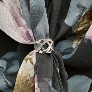 May include: A silver scarf ring with an interwoven design, adorned with small, clear crystals. The ring fastens a floral-patterned scarf in black, grey, and purple tones. The scarf is made of a lightweight material.