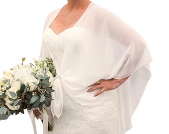 May include: Ivory bridal shawl draped over a white wedding dress. The shawl is tied at the front and has a flowing, lightweight design. A bouquet of white flowers and greenery is held in the hand.