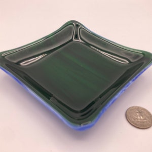 May include: A small, square glass dish with a dark green interior and a light blue rim. The dish is slightly curved and has a glossy finish.