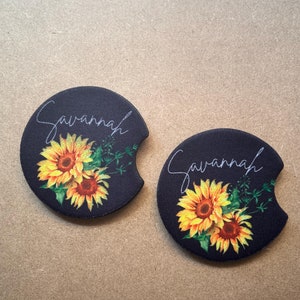 May include: Two black round coasters with a sunflower design and the name "Savannah" written in white script.