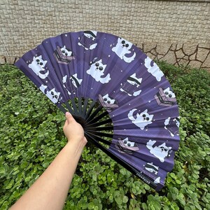 May include: A purple and black hand fan with a white cat design. The fan features a repeating pattern of cats wearing sunglasses and holding a box with the word "CRAVIN'" on it. The fan is open and held by a hand.