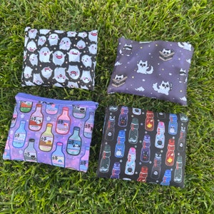 May include: Four reusable shopping bags with different designs. The first bag is white with black ghosts, the second bag is purple with a pattern of soda bottles, the third bag is black with a pattern of white cats, and the fourth bag is black with a pattern of white and black cats.