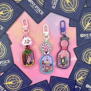 Pu&ograve; includere: Tre portachiavi colorati a forma di bottiglie di vetro con glitter e personaggi diversi all'interno. Le bottiglie sono riempite di glitter e hanno un ciondolo attaccato alla parte superiore. I portachiavi sono rosa, blu e verdi.