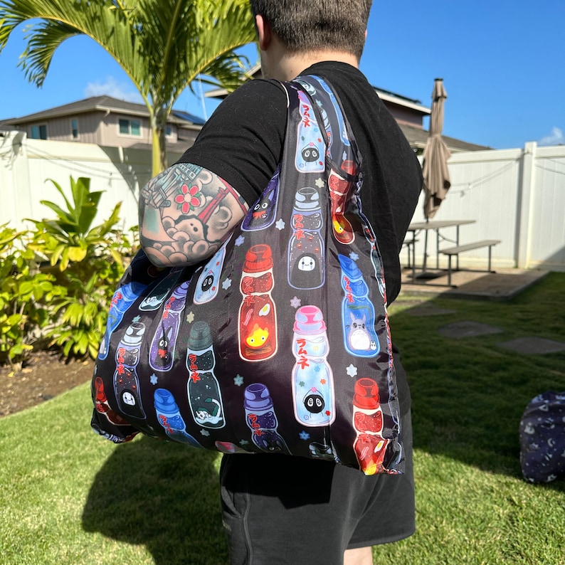 May include: A black reusable tote bag with a colorful print of cartoon characters and Japanese text. The bag is made of a lightweight fabric and has a long strap.