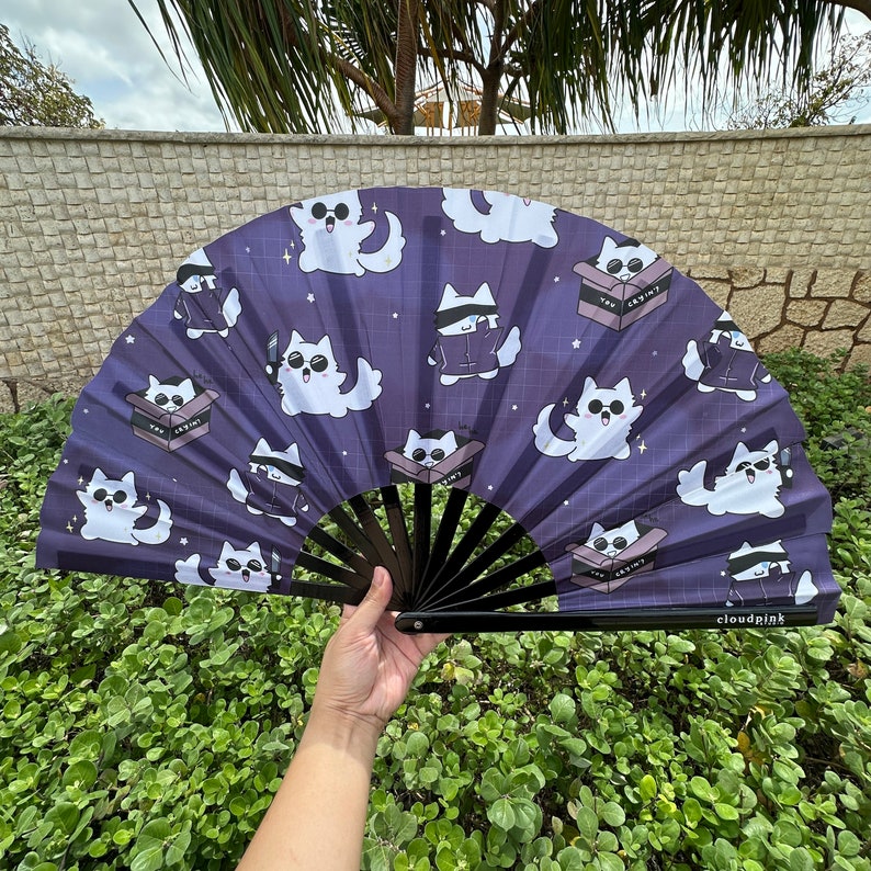 May include: A purple and white hand fan with a black handle. The fan features a repeating pattern of white cartoon cats in various poses. The text "TOY SKINT" appears on the fan.