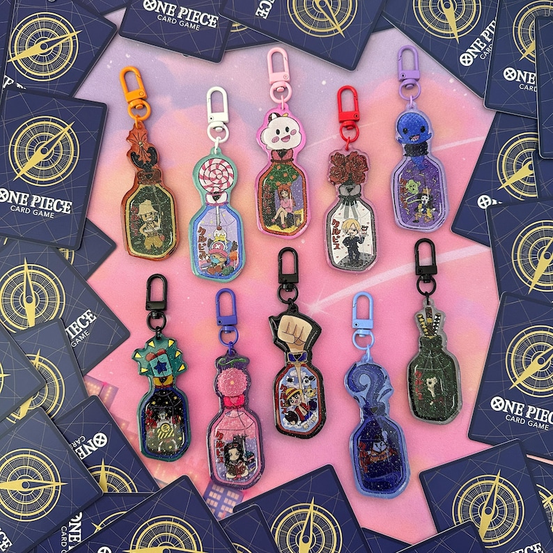 Pu&ograve; includere: Dieci portachiavi colorati con personaggi dell'anime One Piece. Ogni portachiavi &egrave; una piccola bottiglia con glitter e un personaggio all'interno. I portachiavi sono su uno sfondo rosa con carte blu e dorate.