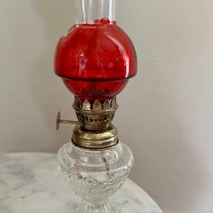 May include: A vintage-style oil lamp featuring a clear glass base with a textured, beaded pattern, a brass-toned metal burner assembly with an adjustment knob, a vibrant red glass globe, and a tall, clear glass chimney extending from the top.