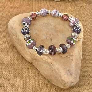 Peut inclure: Un bracelet de perles en verre violet et blanc avec des entretoises argentées. Le bracelet est sur un rocher brun clair.