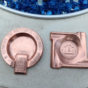 May include: Two vintage copper ashtrays with a design of the Chicago skyline. The larger ashtray is round and reads "A Century of Progress Chicago 1934". The smaller ashtray is square and has a raised design of the Chicago skyline.