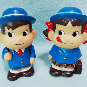 Puede incluir: Dos figuritas con uniforme azul y blanco, una masculina y otra femenina, con detalles rojos. La figurita masculina lleva un sombrero azul y una bolsa. La figurita femenina lleva un sombrero azul con lazos rojos y un maletín marrón.