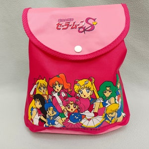 Pode incluir: Uma bolsa rosa com zíper com um gráfico de Sailor Moon S. A bolsa tem uma aba rosa com um fecho de botão branco. O gráfico mostra as Sailor Scouts em suas poses icônicas.