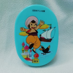 Peut inclure: Un compact ovale bleu avec une image de dessin animé d'un garçon portant un turban et une tenue jaune, sautant en l'air avec un bateau et un oiseau en arrière-plan. Le texte "©日本アニメ企画" est imprimé sur le compact.