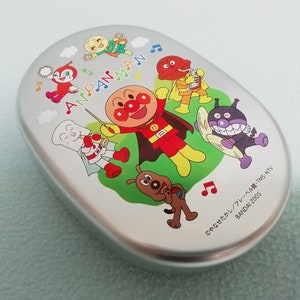 Puede incluir: Una fiambrera de plata con Anpanman y sus amigos. La fiambrera tiene un dise&ntilde;o colorido con Anpanman, un superh&eacute;roe rojo con cabeza de pan, y otros personajes de la serie de anime japonesa. El texto en la fiambrera dice "Anpanman" y "&copy;やなせたかし/フレーベル館・TMS・NTV BANDAI 2005".