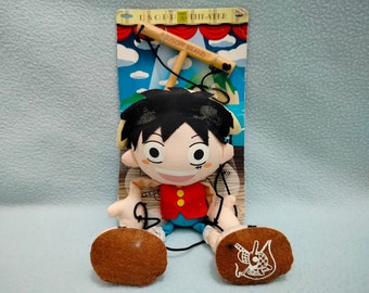 May include: A plush puppet of a cartoon character with black hair, a red shirt, and brown shoes. The puppet is attached to a wooden stick with a string. The packaging features the text "Usopp Theater" and "Usopp Brand".
