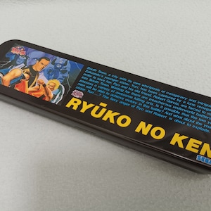 May include: A black metal pencil case featuring a colourful image of characters from the video game "Ryuko no Ken". The case also has the text "Ryuko no Ken" in yellow letters and the Sega logo.
