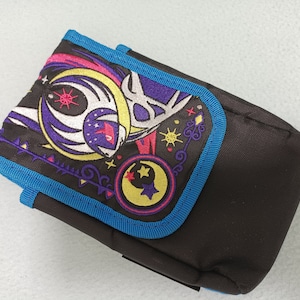 May include: A rectangular, brown fabric pouch with a flap, trimmed in bright blue. The flap features an embroidered design with a crescent moon, stars, and a celestial-themed illustration in purple, yellow, and pink.