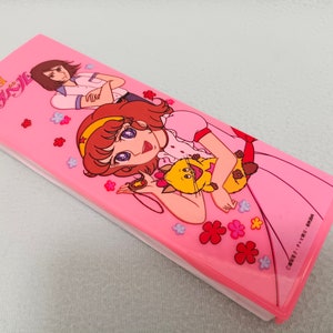 May include: A pink pencil case with a cartoon image of two girls and a cat. The case has the text "魔法の天使 クリスタル" on the front.