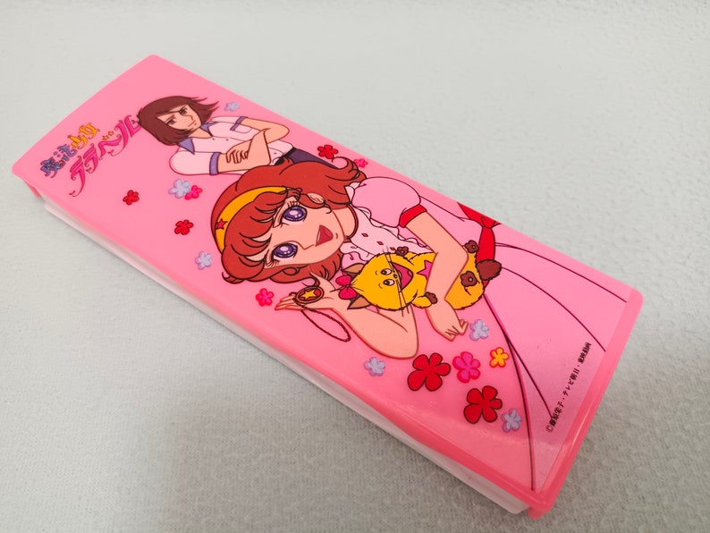 May include: A pink pencil case with a cartoon image of two girls and a cat. The case has the text "魔法の天使 クリスタル" on the front.