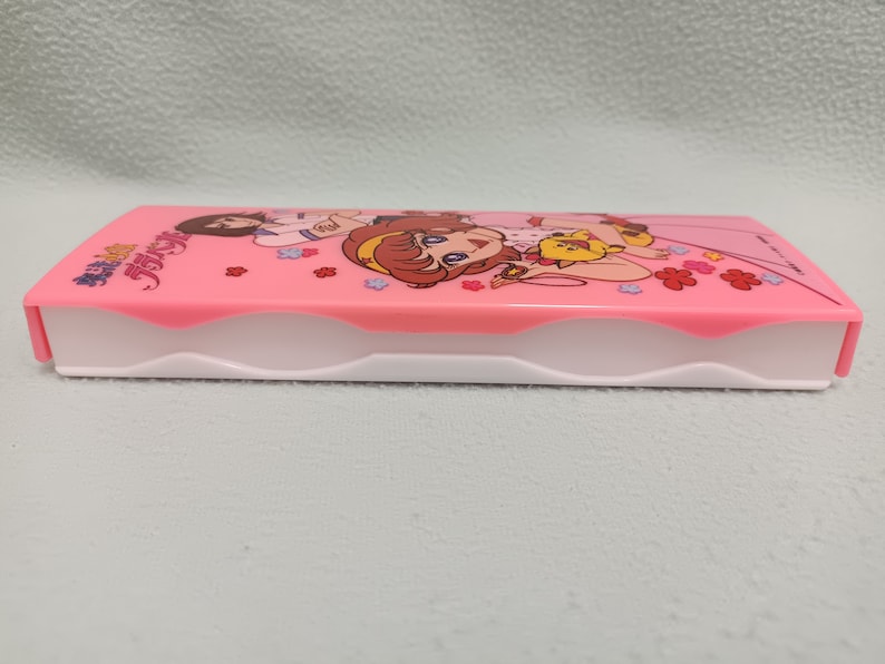 May include: A pink plastic pencil case with a white interior. The case features a cartoon image of two girls with a small animal. The text "魔法少女" is printed on the case.