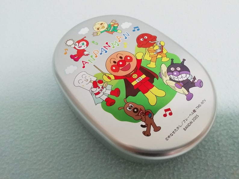 Puede incluir: Una fiambrera de plata con Anpanman y sus amigos. La fiambrera tiene un dise&ntilde;o colorido con Anpanman, un superh&eacute;roe rojo con cabeza de pan, y otros personajes de la serie de anime japonesa. El texto en la fiambrera dice "Anpanman" y "&copy;やなせたかし/フレーベル館・TMS・NTV BANDAI 2005".