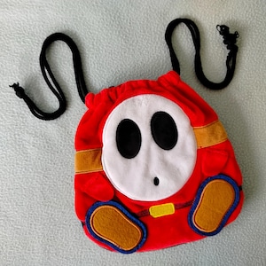 May include: A red plush bag with a Shy Guy character design. The bag has a white face with black eyes, a small nose, and a red body. It includes a black drawstring and gold and blue accents.