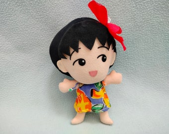 May include: A plush toy of a girl with black hair and a red bow. She is wearing a colorful dress with a floral pattern.