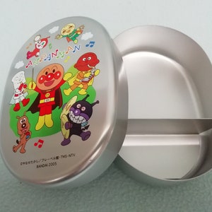 Puede incluir: Una fiambrera de Anpanman plateada con un compartimento dividido. La tapa presenta una ilustraci&oacute;n colorida de Anpanman y sus amigos tocando m&uacute;sica. El texto en la tapa dice "Anpanman" y "BANDAI 2005".