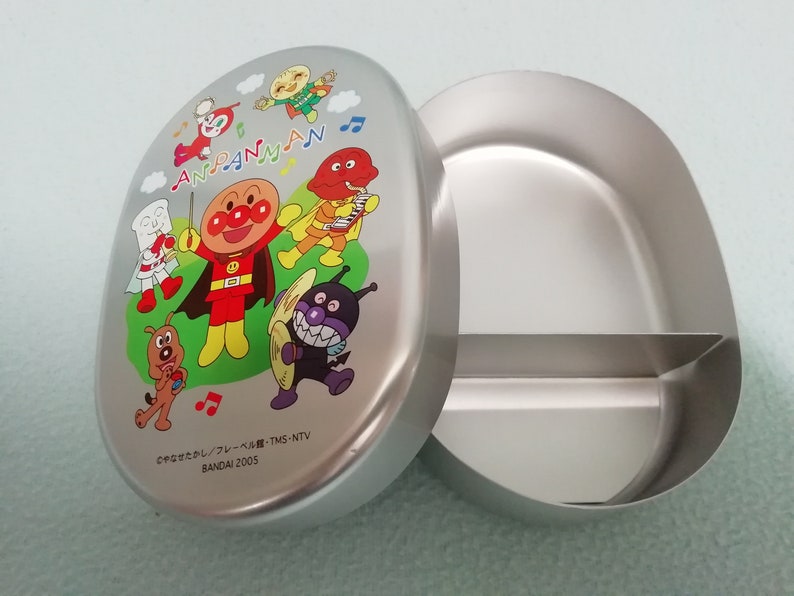 Puede incluir: Una fiambrera de Anpanman plateada con un compartimento dividido. La tapa presenta una ilustraci&oacute;n colorida de Anpanman y sus amigos tocando m&uacute;sica. El texto en la tapa dice "Anpanman" y "BANDAI 2005".