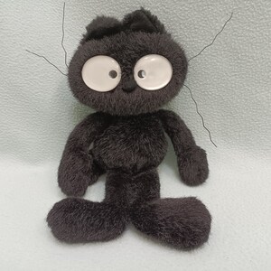 Op de afbeelding: Een zwarte pluche kattenpop met grote witte ogen en lange snorharen. De pop heeft een eenvoudig ontwerp met een kleine neus en mond en staat rechtop. De vacht van de kat is zacht en pluizig.