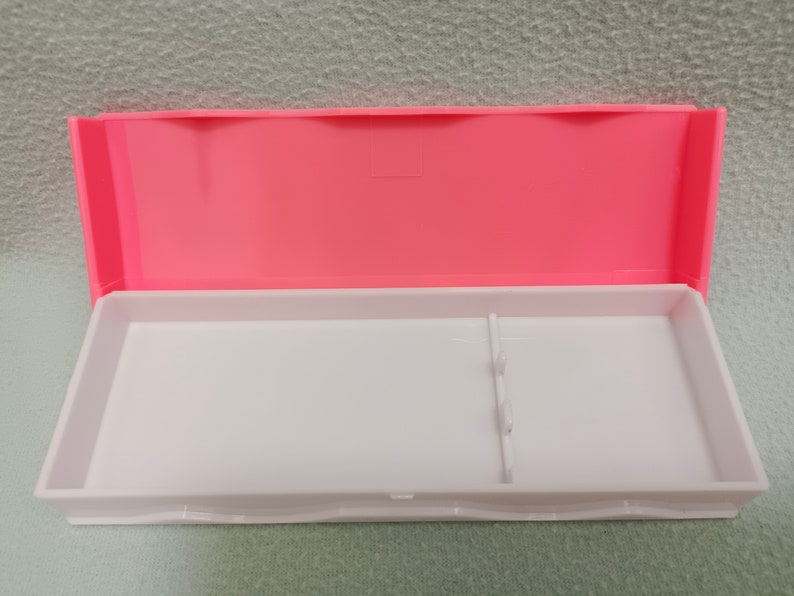 May include: A pink plastic pencil box with a white interior. The box has a divider inside to separate items.