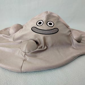 以下が含まれることがあります： 笑顔の顔をしたブロブモンスターの形をした灰色のぬいぐるみ。おもちゃは大きく丸い頭と小さく丸い体を持っています。おもちゃは柔らかく、ぬいぐるみでできており、滑らかでベルベットのような質感です。