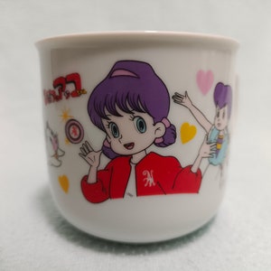 Puede incluir: Taza de cerámica blanca con una ilustración de dibujos animados de una niña con cabello morado que lleva una chaqueta roja. La niña sostiene un objeto rojo y dorado. La taza también presenta una niña de dibujos animados más pequeña con cabello morado que lleva un vestido azul y amarillo. La taza tiene el texto "魔法の天使 クリィミーマミ" en japonés.