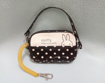 Op de afbeelding: Een zwart-witte polkadot-tas met een wit konijnengezicht en de tekst "miffy Hide-and-seek" aan de voorkant. De tas heeft een gele band met een zilveren sleutelhanger.