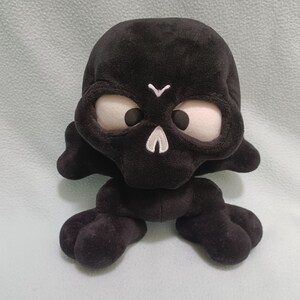May include: A black plush skull toy with large, light-colored eyes and a white stitched design on the forehead and chin. The toy has a soft, velvety texture and is designed with rounded features.